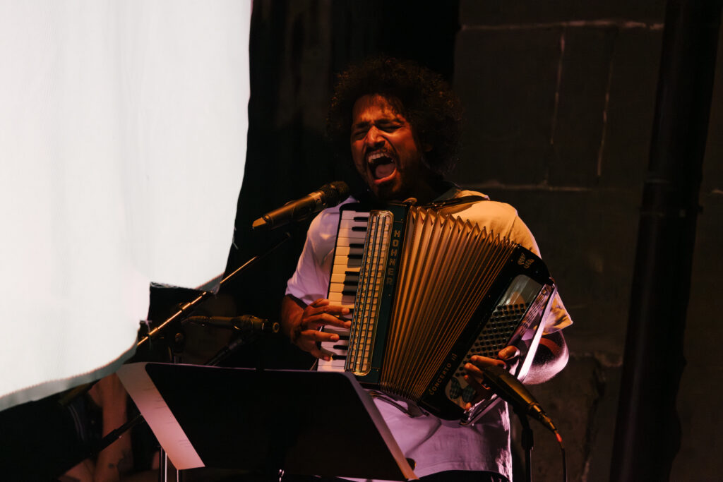 Julian Warner steht auf einer Bühne, singt kraftvoll in ein Mikrofon und spielt ein Hohner‑Concerto‑II‑Akkordeon. Mehrere Mikrofone sind um ihn herum platziert, vor ihm befindet sich ein Notenständer mit Blättern. Die Bühnenbeleuchtung hebt ihn deutlich vom dunklen Hintergrund ab, links ist eine helle Projektionsfläche zu erkennen.