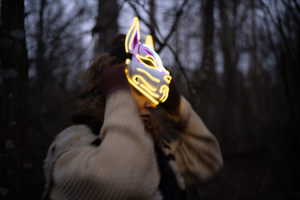 Eine Person steht in einem dunklen Wald zwischen kahlen Bäumen und trägt eine leuchtende Tiermaske mit gelben und violetten Konturen, die an ein Fuchs‑ oder Wolfsgesicht erinnert. Sie hat einen hellen, dicken Pullover und braune Handschuhe an und hält die Maske mit beiden Händen fest.