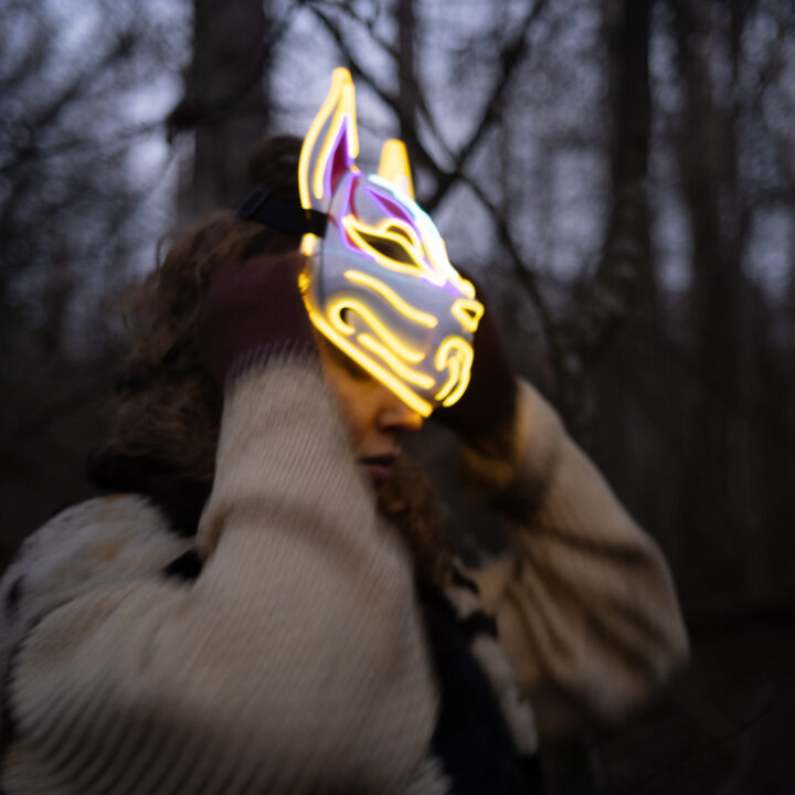 Eine Person steht in einem dunklen Wald zwischen kahlen Bäumen und trägt eine leuchtende Tiermaske mit gelben und violetten Konturen, die an ein Fuchs‑ oder Wolfsgesicht erinnert. Sie hat einen hellen, dicken Pullover und braune Handschuhe an und hält die Maske mit beiden Händen fest.