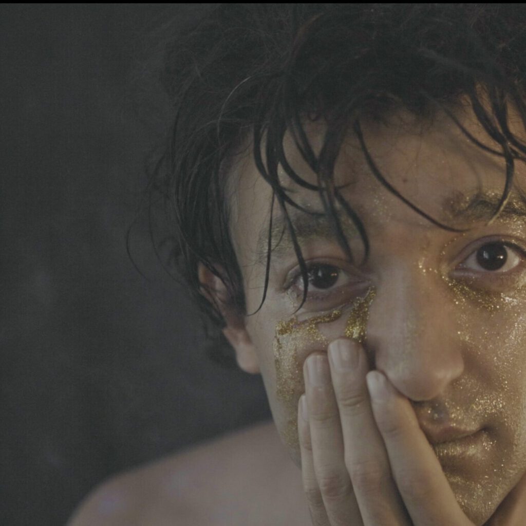 Eine Person mit nassem, dunklem Haar ist in einer Nahaufnahme zu sehen. Das Gesicht ist großflächig mit goldfarbenem Glitter oder Metallic‑Pigment bedeckt, besonders um Augen, Stirn und Wangen. Eine Hand verdeckt teilweise Mund und Kinn, wodurch der Blick auf die schimmernde Textur und den intensiven Ausdruck fokussiert wird. Der Hintergrund ist dunkel und schlicht, was den goldenen Glanz und die intime, fast performative Stimmung der Aufnahme betont.