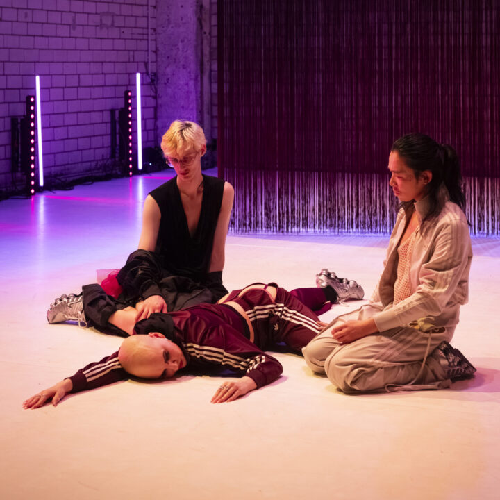 In einem von violett‑rosa Licht durchfluteten Raum liegen und sitzen drei Personen in einer performativen Szene. Eine Person trägt einen weinroten Trainingsanzug mit weißen Streifen und liegt ausgestreckt auf dem Boden. Zwei weitere Personen sitzen daneben, eine in schwarzer Kleidung, die andere in einem hellen Outfit. Hinter ihnen steht eine Backsteinwand, vor der lange, vertikale Fäden oder Stoffbahnen hängen, durch LED‑Lichtstäbe atmosphärisch beleuchtet.