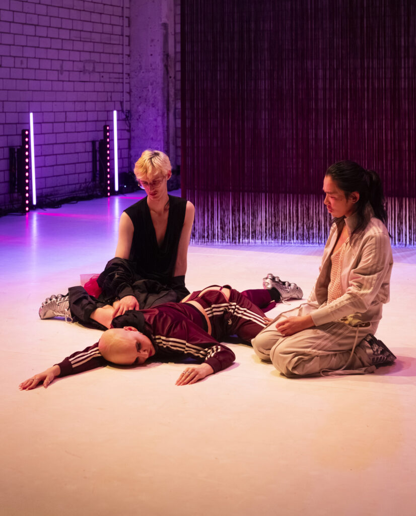 In einem von violett‑rosa Licht durchfluteten Raum liegen und sitzen drei Personen in einer performativen Szene. Eine Person trägt einen weinroten Trainingsanzug mit weißen Streifen und liegt ausgestreckt auf dem Boden. Zwei weitere Personen sitzen daneben, eine in schwarzer Kleidung, die andere in einem hellen Outfit. Hinter ihnen steht eine Backsteinwand, vor der lange, vertikale Fäden oder Stoffbahnen hängen, durch LED‑Lichtstäbe atmosphärisch beleuchtet.