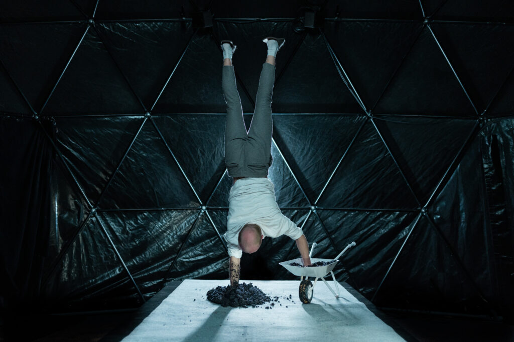 Eine Person steht in einem dunklen, kuppelförmigen Raum aus dreieckigen Paneelen und macht einen Handstand. Sie trägt ein weißes Shirt, graue Hose und weiße Socken. Eine Hand stützt sich in einem Haufen dunkler Erde oder Kies auf einer weißen Fläche, die andere ruht in einer kleinen weißen Schubkarre, die ebenfalls mit Erde gefüllt ist. Das Licht fällt konzentriert auf die Person und die Materialien, wodurch ein starker Kontrast zum dunklen Hintergrund entsteht und die ungewöhnliche, performative Szene betont wird.