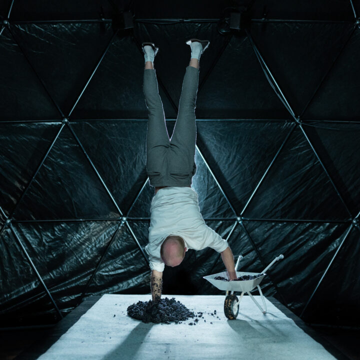 Eine Person steht in einem dunklen, kuppelförmigen Raum aus dreieckigen Paneelen und macht einen Handstand. Sie trägt ein weißes Shirt, graue Hose und weiße Socken. Eine Hand stützt sich in einem Haufen dunkler Erde oder Kies auf einer weißen Fläche, die andere ruht in einer kleinen weißen Schubkarre, die ebenfalls mit Erde gefüllt ist. Das Licht fällt konzentriert auf die Person und die Materialien, wodurch ein starker Kontrast zum dunklen Hintergrund entsteht und die ungewöhnliche, performative Szene betont wird.