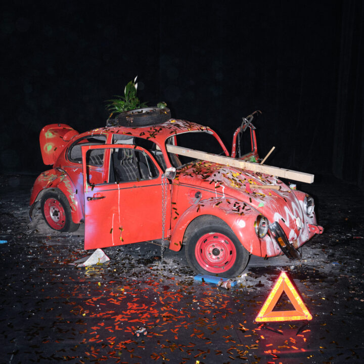 Ein stark beschädigter roter VW‑Käfer steht in einem dunklen Innenraum wie auf einer Bühne. Die Türen sind geöffnet, die Karosserie ist mit buntem Konfetti, Farbspritzern und Kratzern bedeckt. Durch den Bereich der Windschutzscheibe ragt ein Holzbrett, auf dem Dach steht ein Reifen mit einem Blumentopf darin. Vor dem Auto liegt ein rotes Warndreieck, umgeben von verstreutem Metallkonfetti und weiteren kleinen Objekten. Die Szene wirkt wie eine künstlerische Installation, die Zerstörung und Dekoration miteinander verbindet.