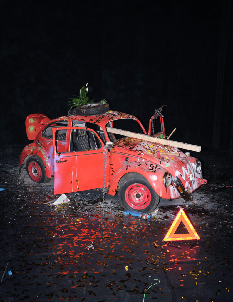 Ein stark beschädigter roter VW‑Käfer steht in einem dunklen Innenraum wie auf einer Bühne. Die Türen sind geöffnet, die Karosserie ist mit buntem Konfetti, Farbspritzern und Kratzern bedeckt. Durch den Bereich der Windschutzscheibe ragt ein Holzbrett, auf dem Dach steht ein Reifen mit einem Blumentopf darin. Vor dem Auto liegt ein rotes Warndreieck, umgeben von verstreutem Metallkonfetti und weiteren kleinen Objekten. Die Szene wirkt wie eine künstlerische Installation, die Zerstörung und Dekoration miteinander verbindet.