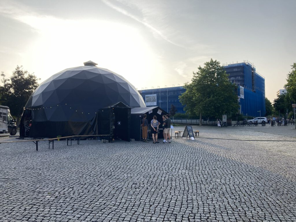Eine große schwarze geodätische Kuppel, die als Kuppelzelt dient, steht auf einem gepflasterten Platz. Vor dem Eingang mit kleinem Vordach und Lichterkette stehen einige Menschen, daneben Bänke und ein Aufsteller. Im Hintergrund sind Bäume und Gebäude mit blauer Bauvernetzung im warmen Abendlicht.